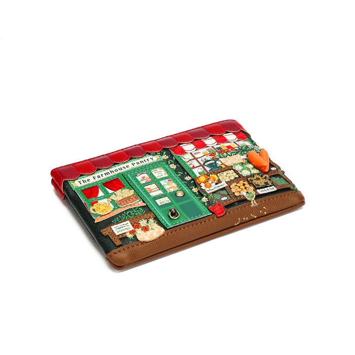 A small brown faux-leather wallet featuring a highly detailed, colorful cartoon illustration of 'The Farmhouse Cafe' on the front. The wallet has a red awning, green storefront, and various whimsical vegetable-themed accents, including a small orange carrot and red tomatoes.