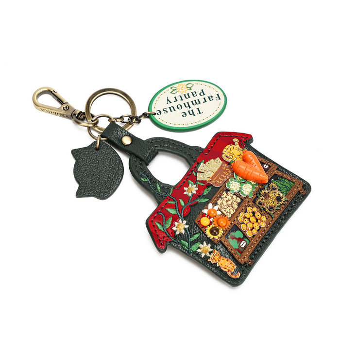 A decorative keychain featuring a handcrafted, rustic house-shaped charm with embroidered floral details and raised vegetable motifs (carrots, peas, beans, corn). The keychain includes a brass clasp, a dark green textured leather clover charm, and a green circular tag reading 'The Farmhouse Parity'.