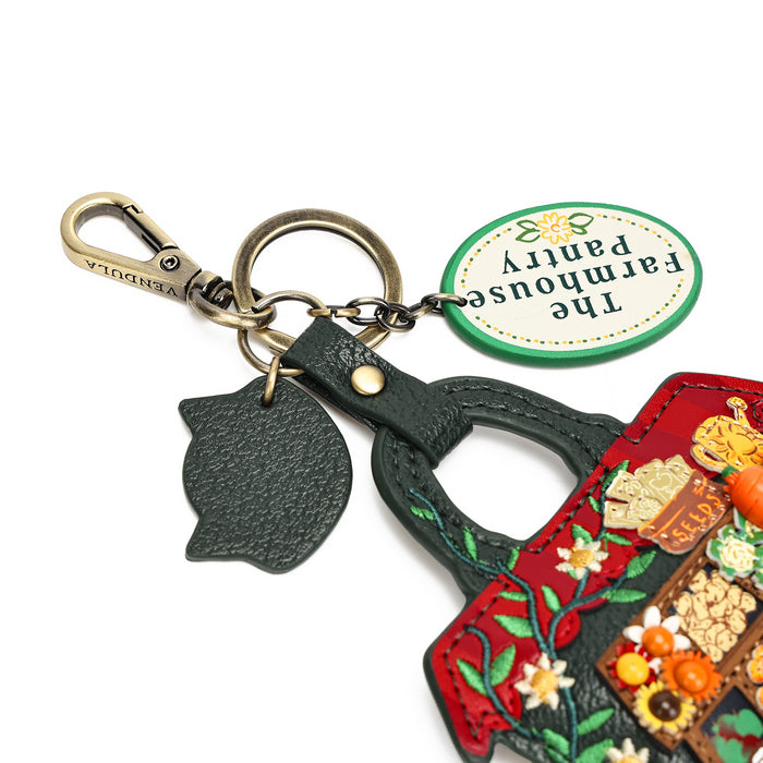 A close-up of a decorative keychain set on a white background, featuring a brass carabiner, a dark green textured key ring with a matching keychain loop and leaf-shaped charm, a round green tag reading 'The Farmhouse Pantry,' and part of a red embroidered purse charm with floral details.