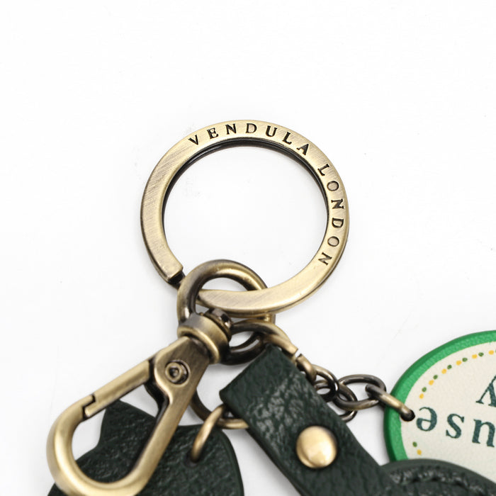 Close-up of a metal key ring set with a brass-toned key ring engraved with 'VENDULA LONDON', a brass-toned lobster clasp, dark green leather strap, and part of a round tag on a white background.