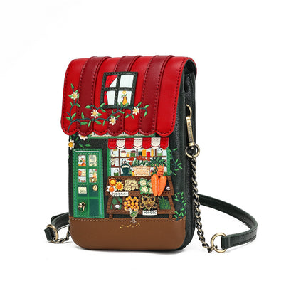 A small crossbody bag with a detailed, colorful illustration of a market stall on the front flap. The bag features a red faux-leather awning with embroidered flowers, a green door, a detailed shop window scene, and a brown base, complete with a black leather strap and a gold-tone chain link.