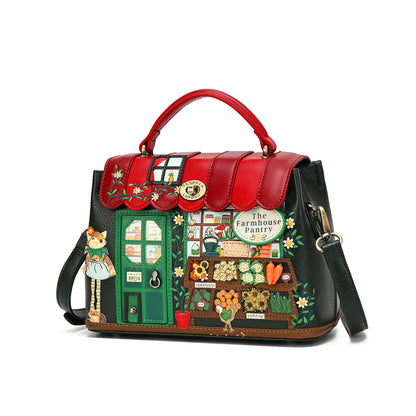 A decorative handbag with a red leather top handle and flap, black leather sides, and a whimsical embroidered storefront patchwork design on the front featuring a green door, flowers, vegetables, and the text 'The Farmhouse Pantry.'.