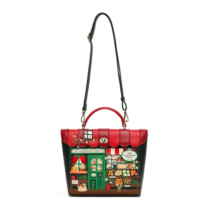 A structured handbag designed to look like 'The Freshhouse Bakery' from The Freshhouse Mystery Shop, featuring a red and black exterior with a detailed bakery storefront illustration on the front, complete with a green door, red awning, and colorful window displays. It has a top handle, a long shoulder strap, and gold-tone hardware.