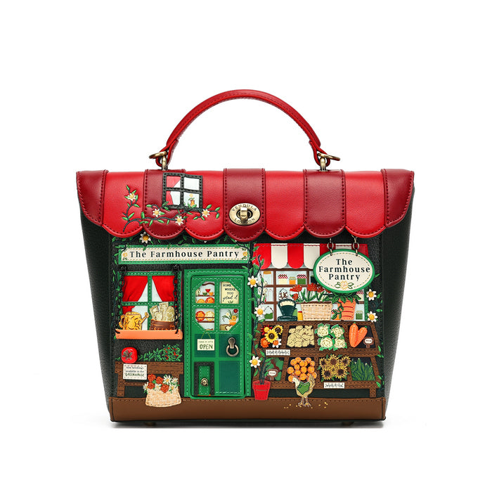 A structured handbag with a red top handle and a front panel printed with a whimsical farm market scene under a red awning, reading 'The Farmhouse Pantry.' The bag has red leather trim, a gold-tone oval clasp, and dark side panels, photographed on a plain white background.