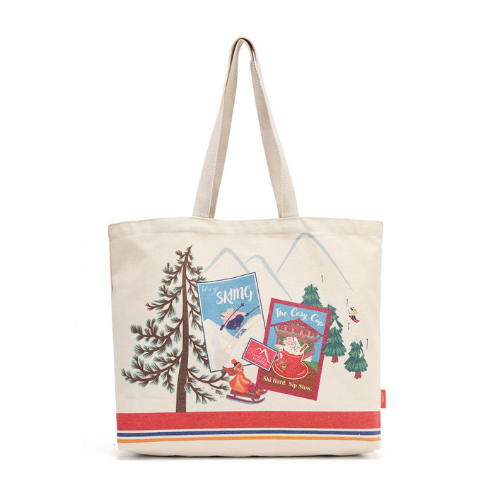 A cream-colored canvas tote bag featuring a winter-themed illustration with a pine tree, mountains, a skier, a hot cocoa cup, and a red stripe at the bottom.