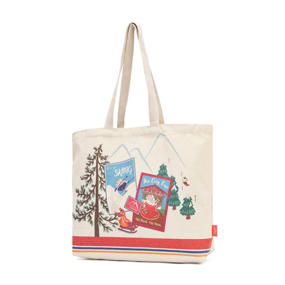 An off-white canvas tote bag with beige shoulder straps, featuring a colorful winter-themed graphic print with mountains, trees, a sleigh, and red and blue text panels, finished with a red, white, and blue stripe along the bottom.