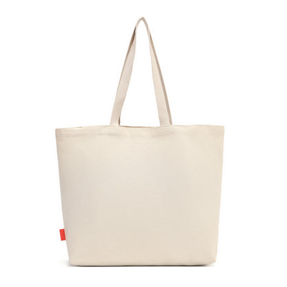 A plain canvas tote bag in a light cream or off-white color, featuring two long shoulder straps and a small red fabric tag on the lower left side, set against a solid white background.