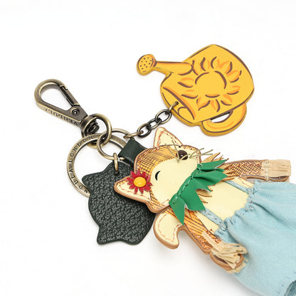 A wooden keychain charm featuring a cute cartoon fox wearing a green scarf and blue skirt, with a gold watering can keychain and a black textured leaf keychain on a white background.