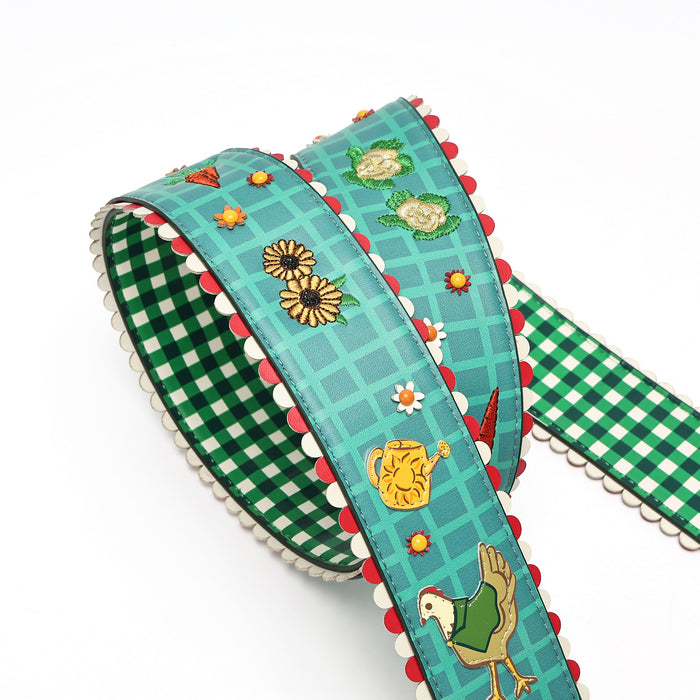 A decorative ribbon or strap with a green and white gingham pattern, turquoise blue grid background, scalloped red and white edges, and embroidered-style motifs including flowers, a watering can, and a chicken, shown on a white background.