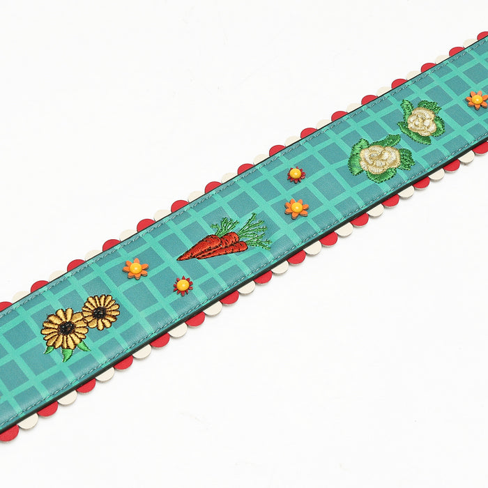 A decorative embroidered ribbon trim with a teal grid pattern, featuring embroidered carrots, daisies, and flowers, finished with a red and white scalloped edge.