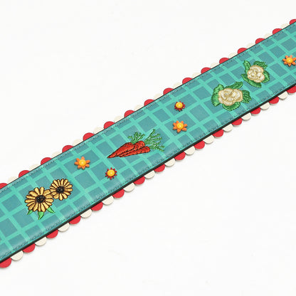 A decorative embroidered ribbon trim with a teal grid pattern, featuring embroidered carrots, daisies, and flowers, finished with a red and white scalloped edge.