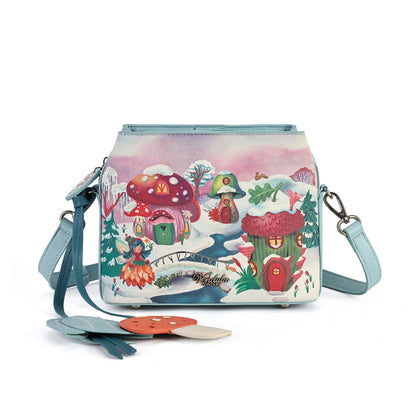 A square-shaped light blue crossbody handbag featuring a whimsical winter forest illustration with mushroom houses, a snowy bridge, and a small treehouse. The bag has a matching light blue strap and silver-tone hardware, with decorative tag accents hanging from the side.