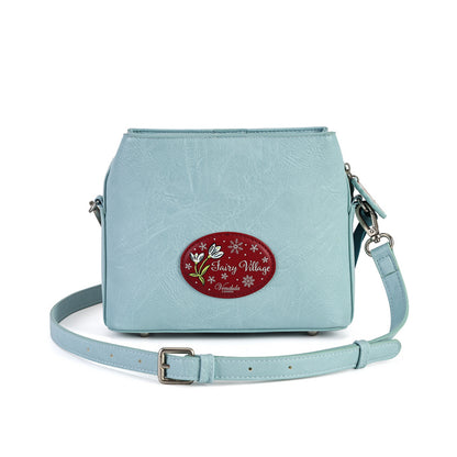 A light blue faux-leather crossbody bag with a decorative red oval patch on the front that reads 'Hazy Village Handmade' with white flowers and snowflakes. The bag has a matching light blue adjustable shoulder strap with a silver buckle, set against a plain white background.