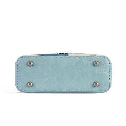 A square-shaped light blue crossbody handbag featuring a whimsical winter forest illustration with mushroom houses, a snowy bridge, and a small treehouse. The bag has a matching light blue strap and silver-tone hardware, with decorative tag accents hanging from the side.