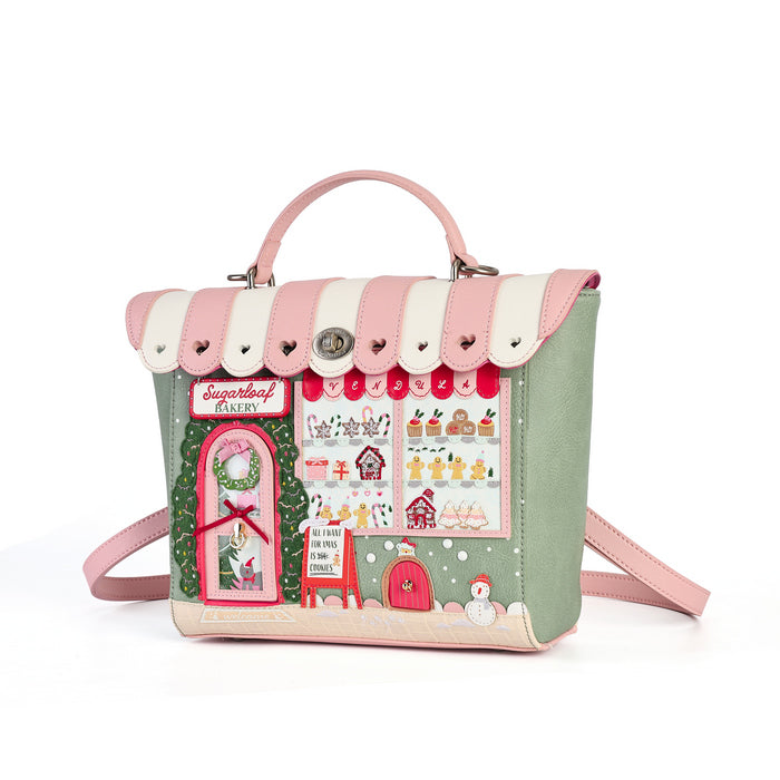 A pastel pink and sage green handbag designed to look like a whimsical storefront or house facade, featuring a pink top handle, adjustable shoulder strap, and a detailed holiday-themed front panel with illustrated windows, a red door, and festive decorations.