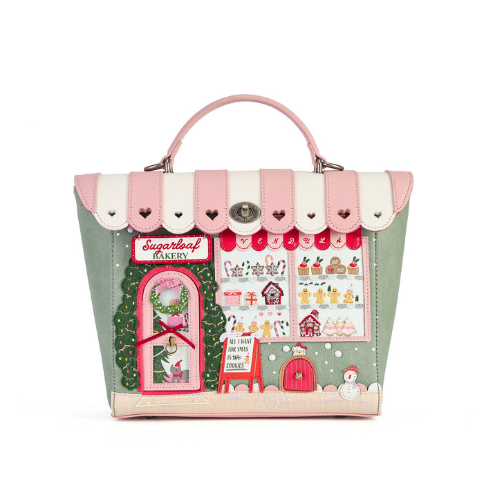A structured handbag with a pink and cream striped top handle and flap, featuring a detailed Christmas bakery storefront applique on the front with green, red, pink, and white elements.
