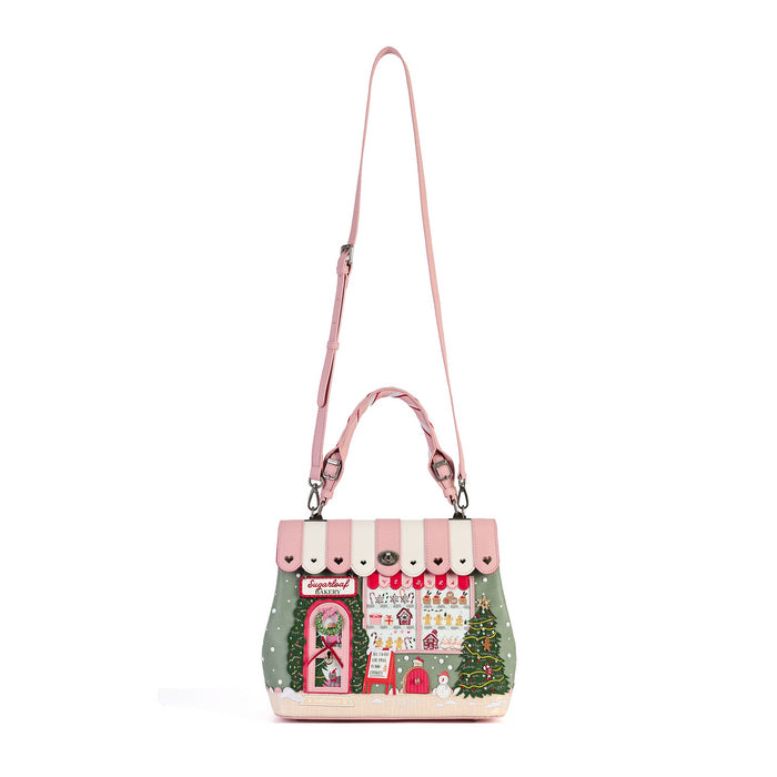 A pink and white striped handbag with a whimsical Christmas village scene printed on the front, featuring a green side panel with a tree, a pink door, and a long shoulder strap, shown on a plain white background.