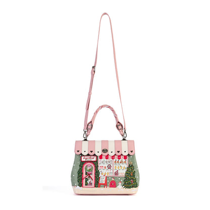 A pink and white striped handbag with a whimsical Christmas village scene printed on the front, featuring a green side panel with a tree, a pink door, and a long shoulder strap, shown on a plain white background.