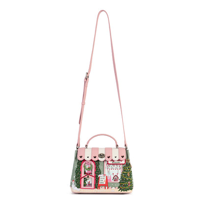 A pink handbag with a holiday village scene patchwork design, featuring a pink shoulder strap, top handle, and embroidered Christmas-themed details including a green tree, red house, and white shop facade on a plain white background.