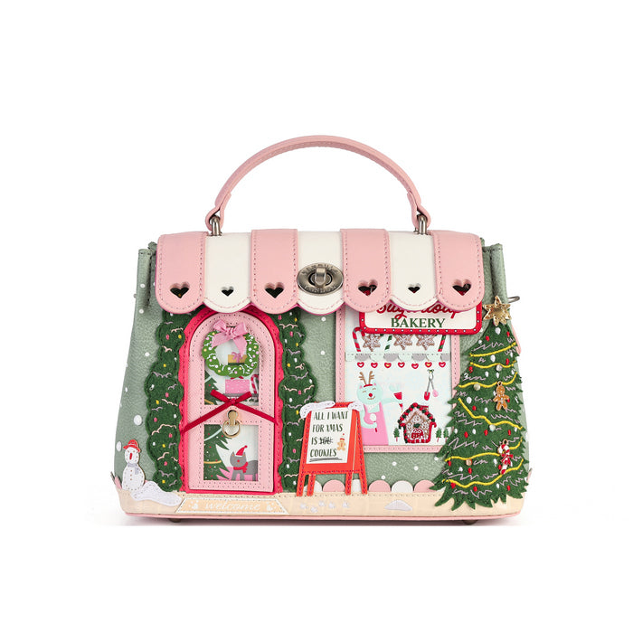 A pink and cream handbag designed to look like a Christmas bakery storefront, featuring a pink top handle, scalloped flap with heart details, a pink window with a wreath, a bakery sign, and Christmas tree illustrations on a white background.