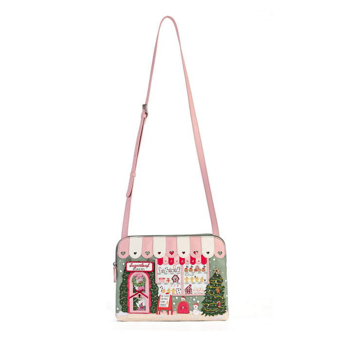 A small shoulder bag with a pink strap, featuring a whimsical Christmas village storefront illustration on the front with pink and white awnings, stockings, a gift box, and a Christmas tree.