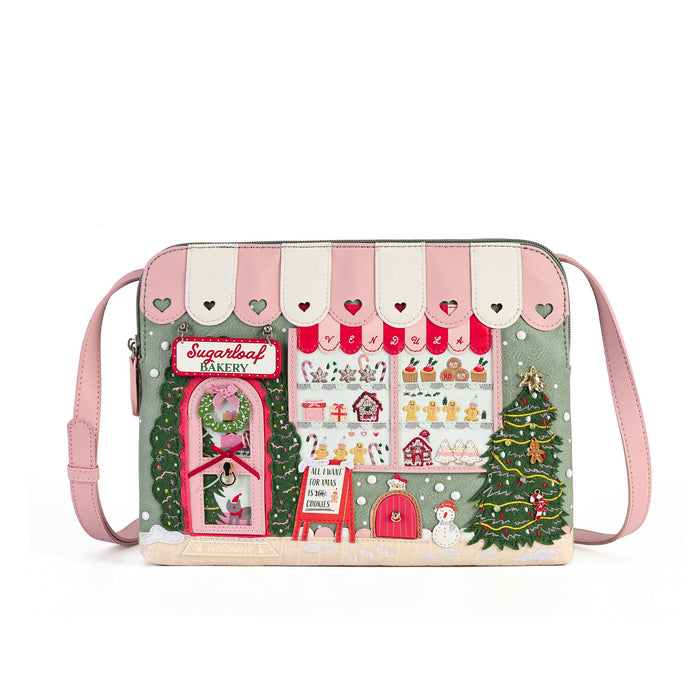 A pastel crossbody bag featuring a whimsical Christmas bakery storefront design. The bag has a light pink strap, a sage green body, and a detailed illustration of a bakery window with a snowman, Christmas trees, and pastries under pink and white awnings.