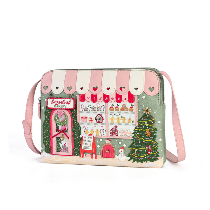 A small shoulder bag with a whimsical Christmas bakery storefront design, featuring pink and cream striped awning, illustrated shop window, holiday tree, and pink strap on a white background.