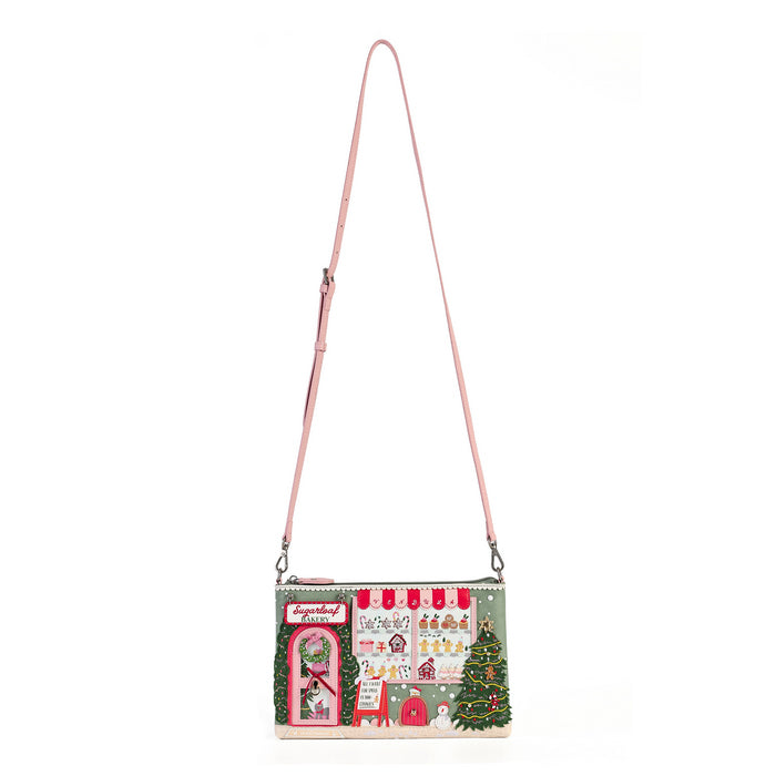 A small shoulder bag with a pink strap, featuring a detailed Christmas village scene printed on the front with green trees, red roofs, and white snow details, set against a plain white background.