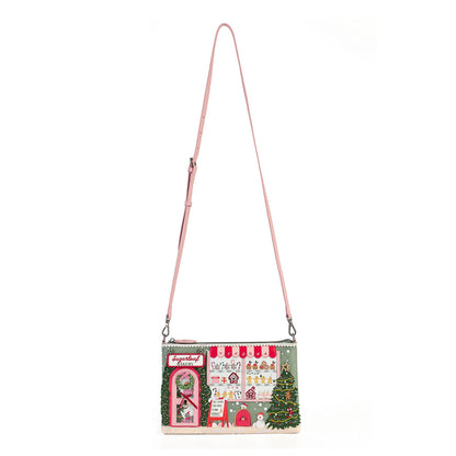A small shoulder bag with a pink strap, featuring a detailed Christmas village scene printed on the front with green trees, red roofs, and white snow details, set against a plain white background.