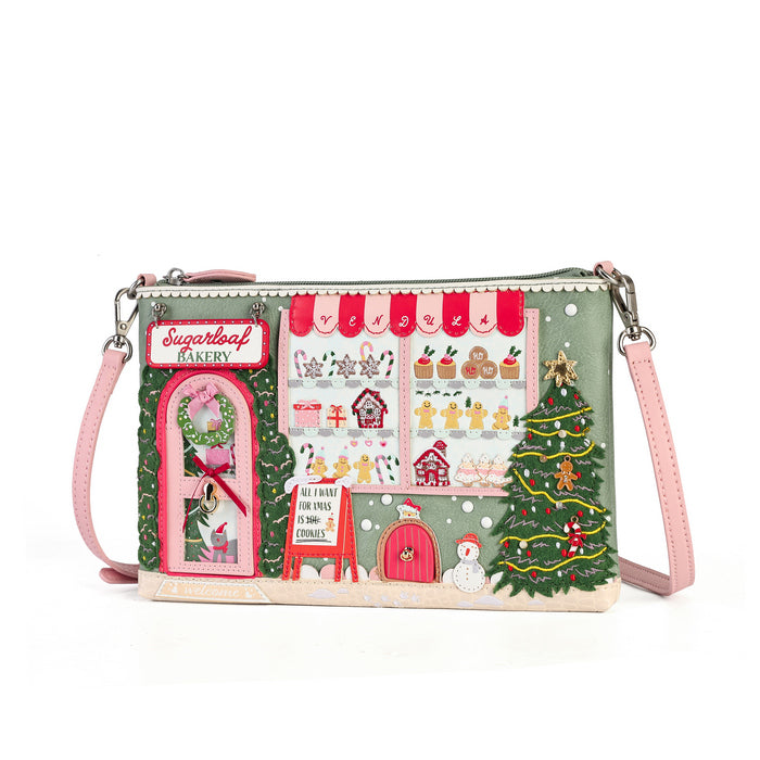 A rectangular crossbody clutch bag with a detailed Christmas bakery scene printed on the front. The design features a green background with red awnings, a pink bakery door labeled 'Sugarleaf Bakery', gingerbread cookies in the window, a decorated Christmas tree, and a snowman.