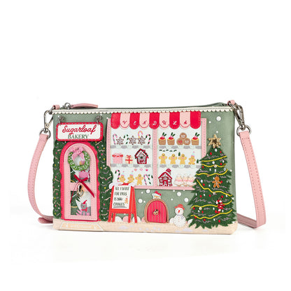 A rectangular crossbody clutch bag with a detailed Christmas bakery scene printed on the front. The design features a green background with red awnings, a pink bakery door labeled 'Sugarleaf Bakery', gingerbread cookies in the window, a decorated Christmas tree, and a snowman.