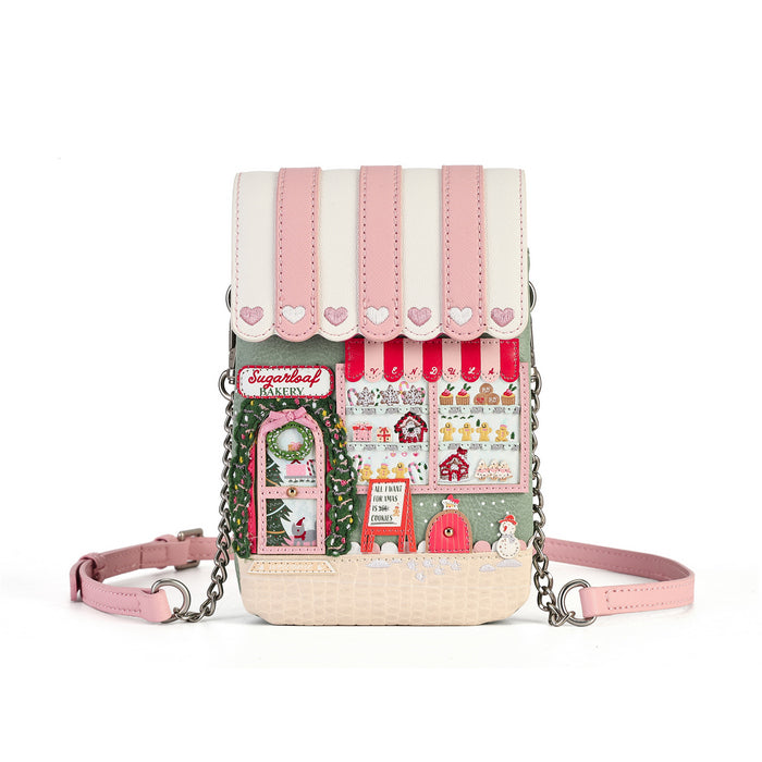 A small crossbody phone bag with a whimsical Christmas village theme, featuring pink and white faux-leather flap panels, a printed front panel with holiday illustrations, a pink strap, and silver chain links, shown on a white background.