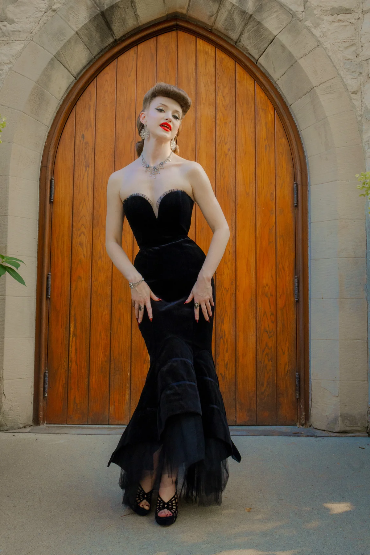 A woman stands in front of a large wooden arched door wearing a black strapless velvet gown with a sweetheart bodice, fitted mermaid-style skirt, and layered tulle hem, paired with black studded heels.