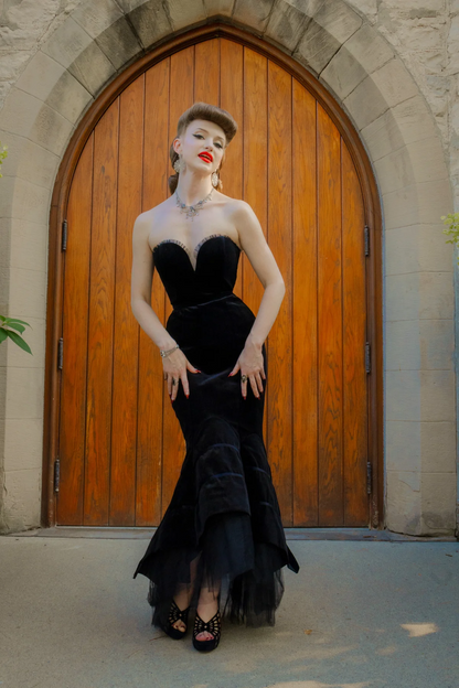 A woman stands in front of a large wooden arched door wearing a black strapless velvet gown with a sweetheart bodice, fitted mermaid-style skirt, and layered tulle hem, paired with black studded heels.