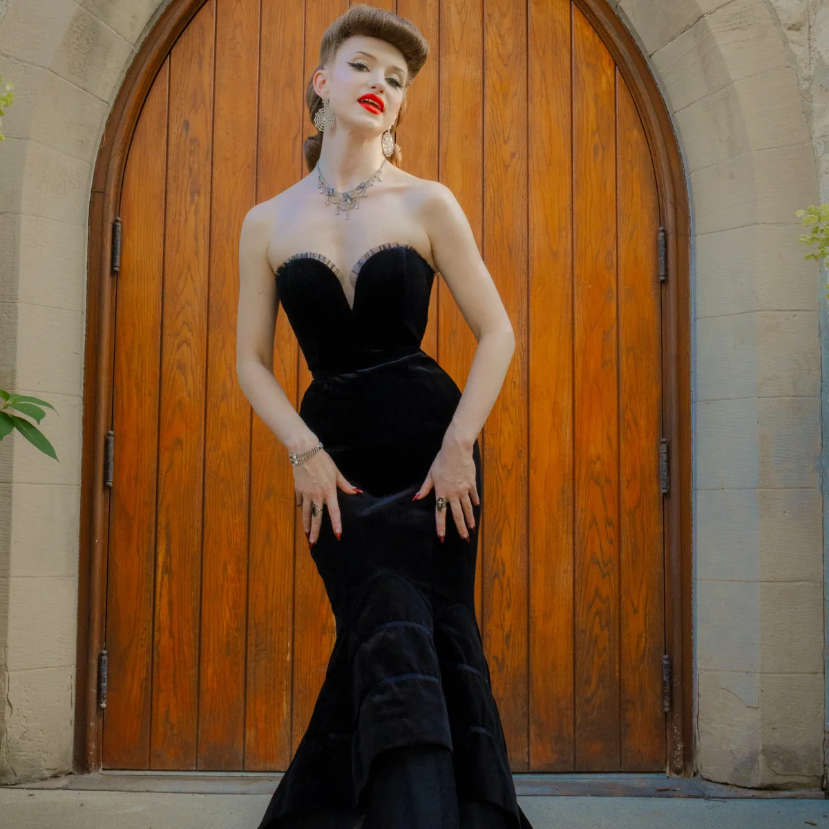 A woman stands in front of a large wooden arched door wearing a black strapless velvet gown with a sweetheart bodice, fitted mermaid-style skirt, and layered tulle hem, paired with black studded heels.
