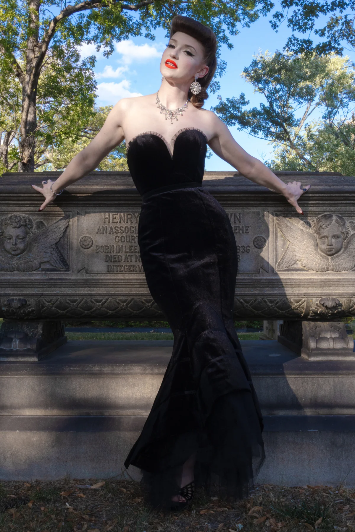 A woman in a black strapless velvet mermaid gown poses outdoors with her arms on a stone bench, with trees and blue sky in the background.
