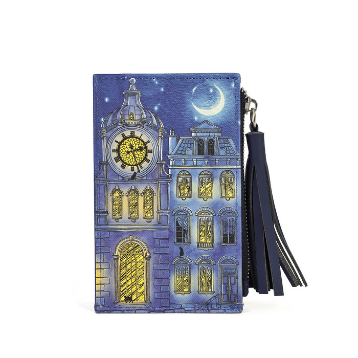 A vertical zip-around wallet featuring a detailed illustration of a whimsical clock tower building at night under a crescent moon. The wallet is decorated with blue and purple tones, glowing yellow windows, and a black leather tassel zipper pull.