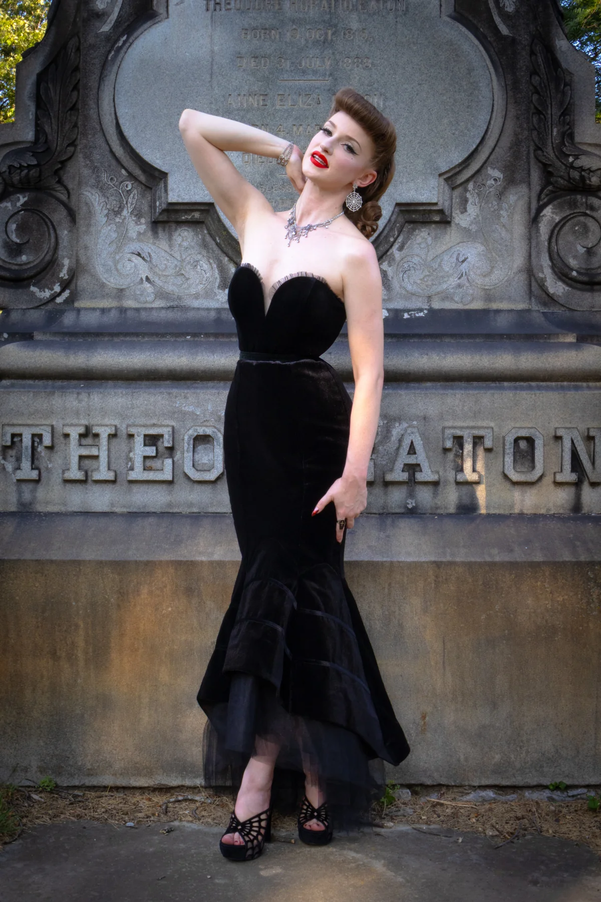A woman posing in a strapless black velvet mermaid gown with a tulle overskirt, standing in front of a large stone monument.