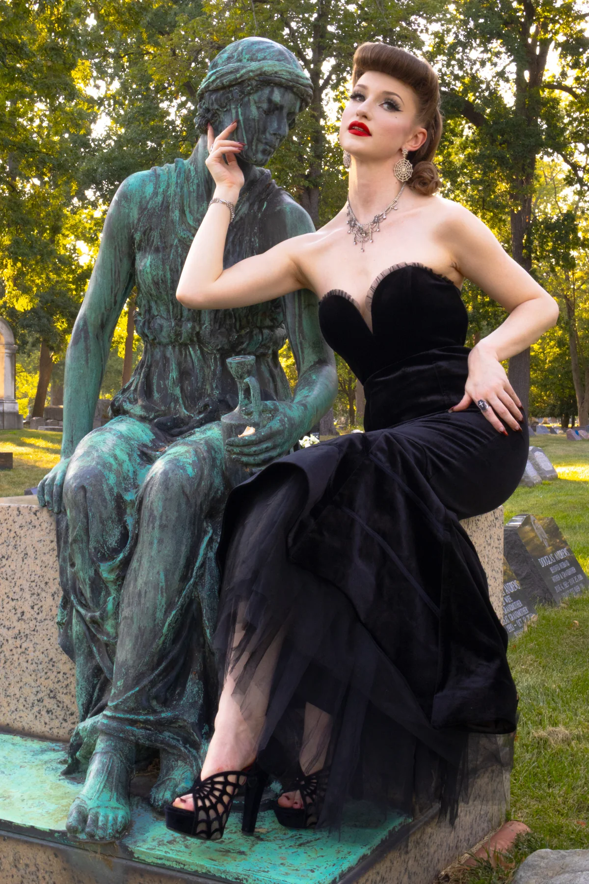 A woman in a black strapless evening gown and black strappy heels sits on a green patina statue in an outdoor park setting.