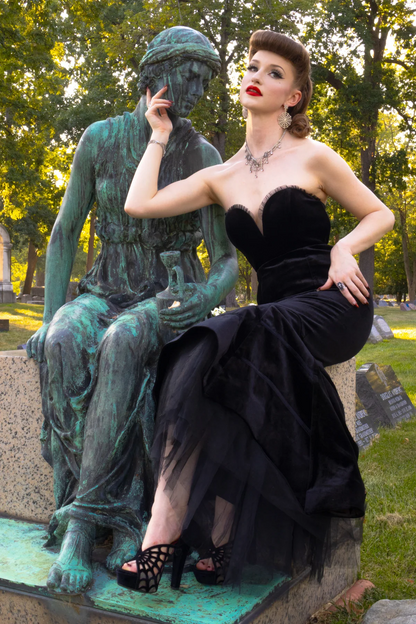A woman in a black strapless evening gown and black strappy heels sits on a green patina statue in an outdoor park setting.