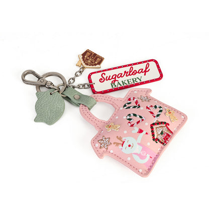 A pink Christmas-themed handbag-shaped keychain bag with a silver clasp, a green leaf charm, a brown bakery tag charm, and a red-and-white 'Sugarloaf Bakery' label. The bag features holiday illustrations including a snowman, gingerbread house, candy canes, and stars on a white background.