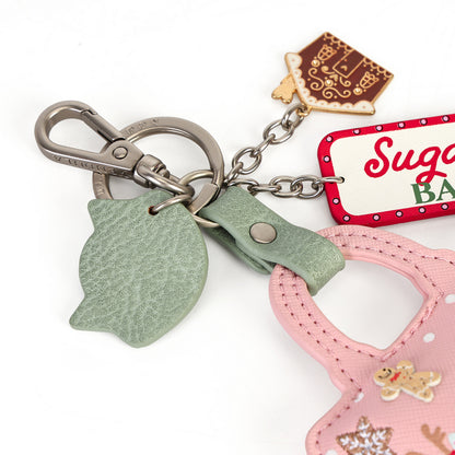 A close-up of a Christmas-themed keychain featuring a silver clasp, a textured sage green leather ornament, a pink fabric bag charm, a red and white 'Sugarcade' tag, and gingerbread cookie charms on a white background.