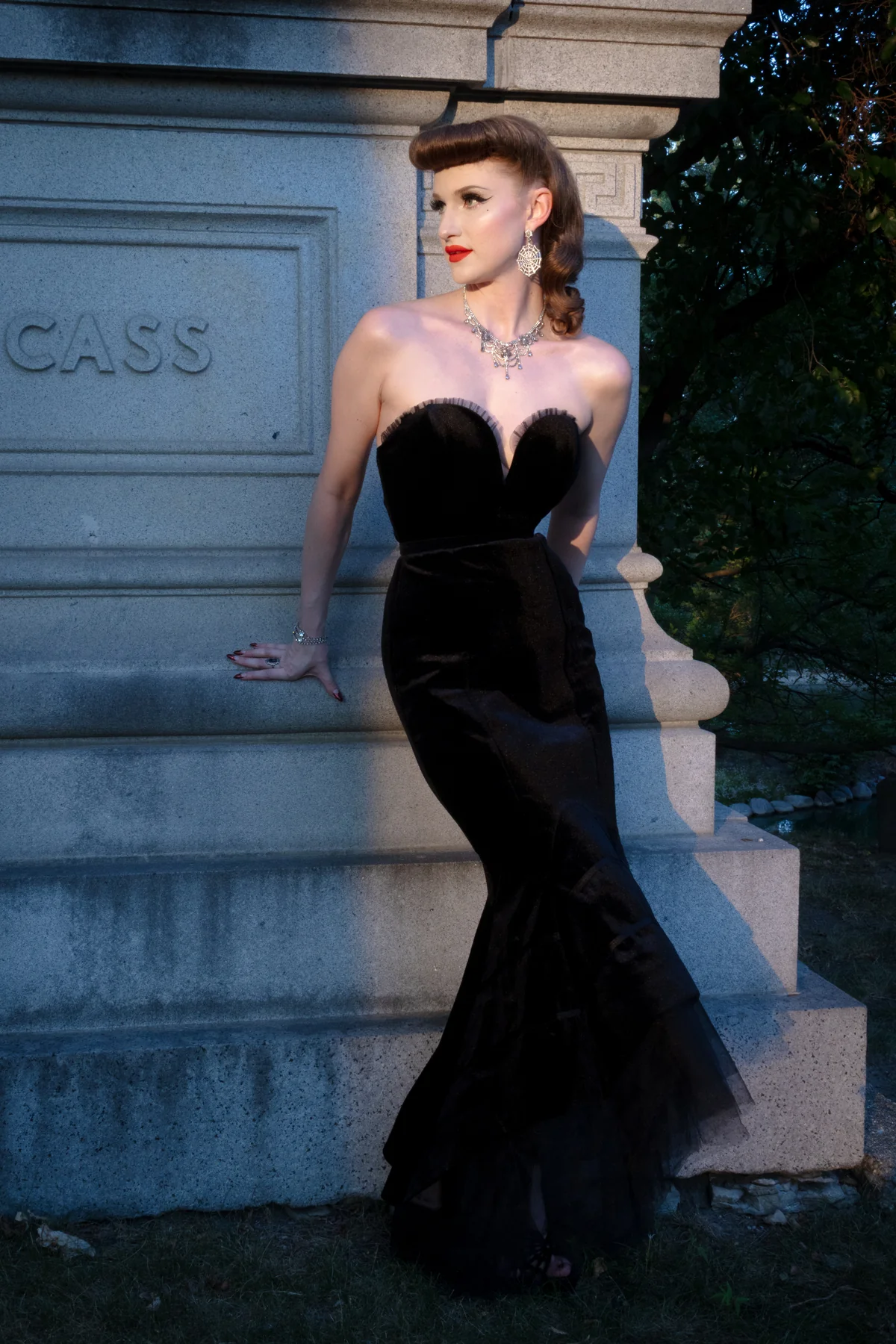 A woman in a black strapless velvet mermaid gown with a sweetheart neckline and flared skirt, posing outdoors against a stone monument.