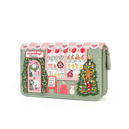 A light sage green zip-around clutch wallet decorated with a detailed Christmas bakery scene. The design features pink and white striped awning, a green wreath door, a bakery window display with pastries, a gingerbread man, a snowman, and a decorated Christmas tree.