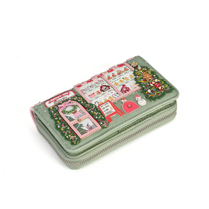 A light sage green zip-around wallet decorated with a colorful Christmas village scene patchwork design, featuring a red postbox, green trees, and festive details, shown on a plain white background.
