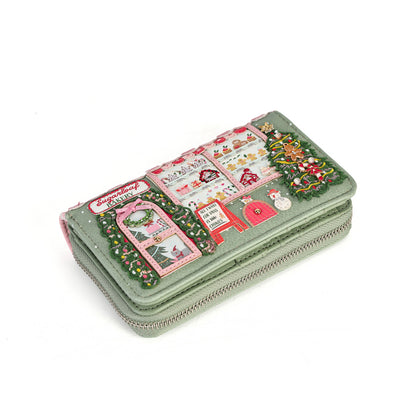 A light sage green zip-around wallet decorated with a colorful Christmas village scene patchwork design, featuring a red postbox, green trees, and festive details, shown on a plain white background.