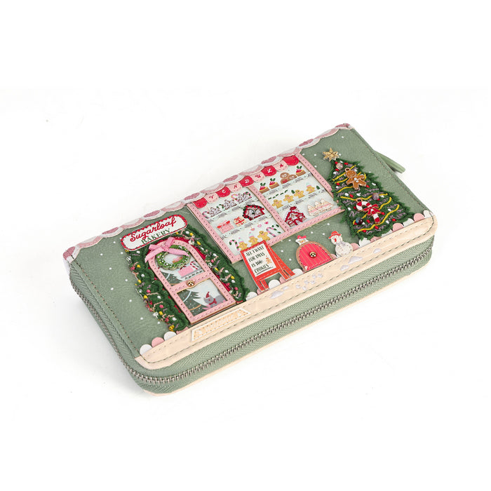 A long zip-around wallet with a festive Christmas village scene printed on the front, featuring a decorated tree, Santa's house, and snowflakes on a muted sage green background.