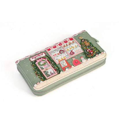 A long zip-around wallet with a festive Christmas village scene printed on the front, featuring a decorated tree, Santa's house, and snowflakes on a muted sage green background.