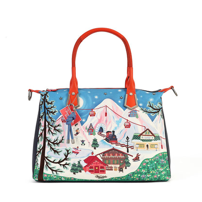 A structured tote bag featuring a colorful winter mountain village illustration on the front panel, with bright red handles and side hardware, photographed against a plain white background.