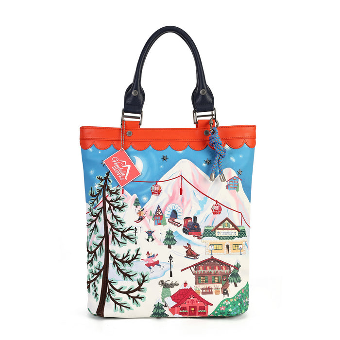A tote bag featuring a colorful winter ski resort illustration on the front, with an orange-red scalloped top border, black leather handles, and a blue plush keychain hanging from the handle.